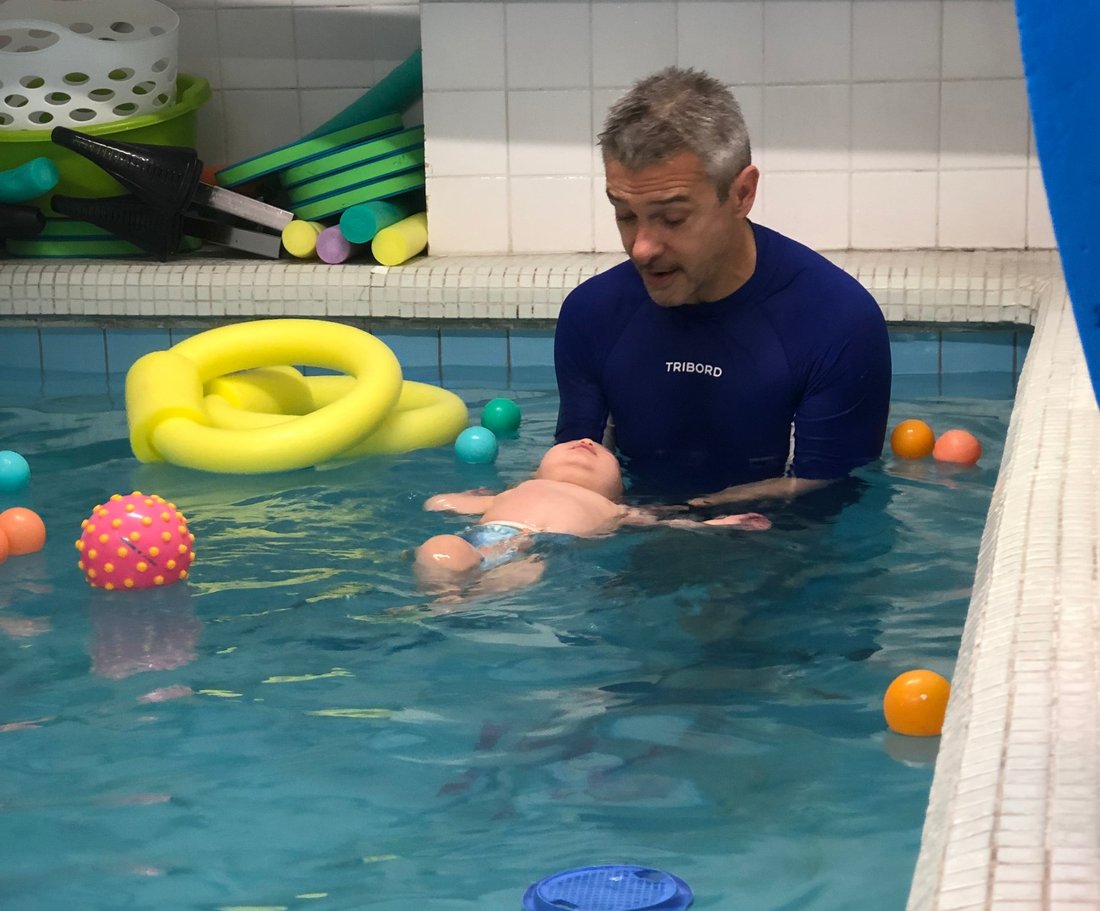 Adrien dans le bassin, soutenant un bébé nageur qui flotte sur le dos au milieu des jouets